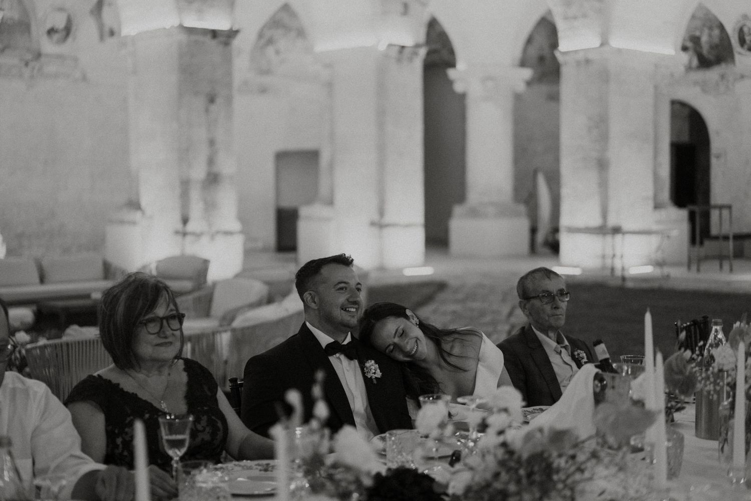 Black and white candid photo of bride leaning on groom at wedding reception dinner table with guests.