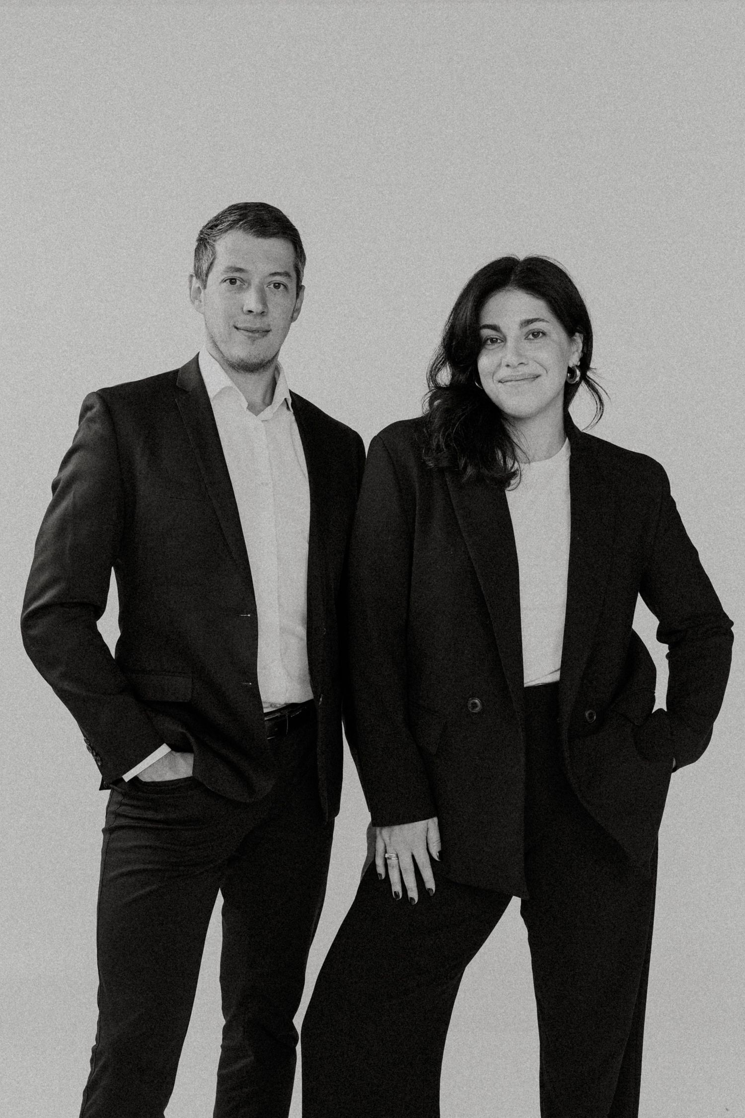 A man and woman in matching black suits pose together for a professional black and white portrait photo.