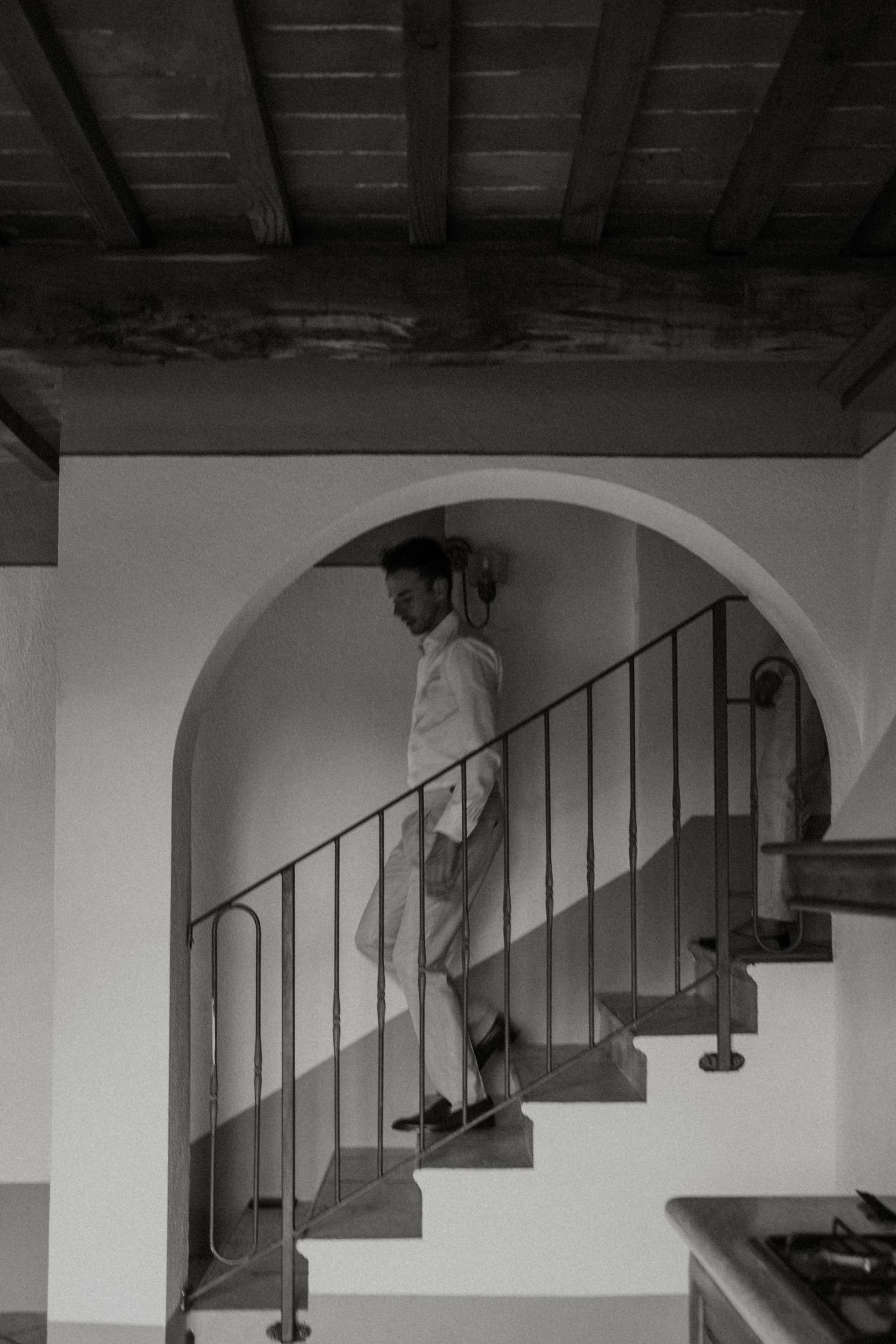 Black and white photo of groom descending rustic staircase under stone arch in Tuscany villa.
