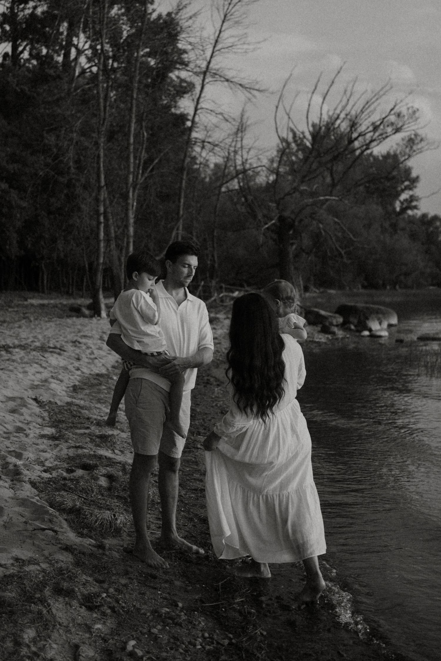 Black and white photo of a family standing by a peaceful river, surrounded by trees, with a woman in a white dress.