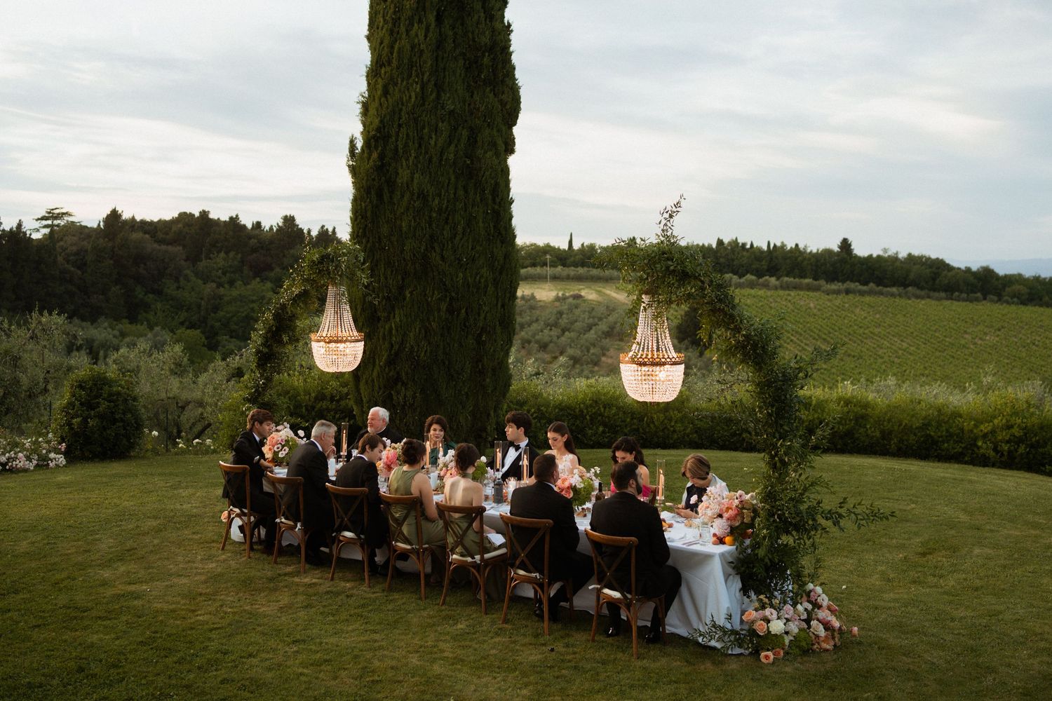 Guests dine at long outdoor table under cypress tree with hanging chandeliers at Tuscan wedding.
