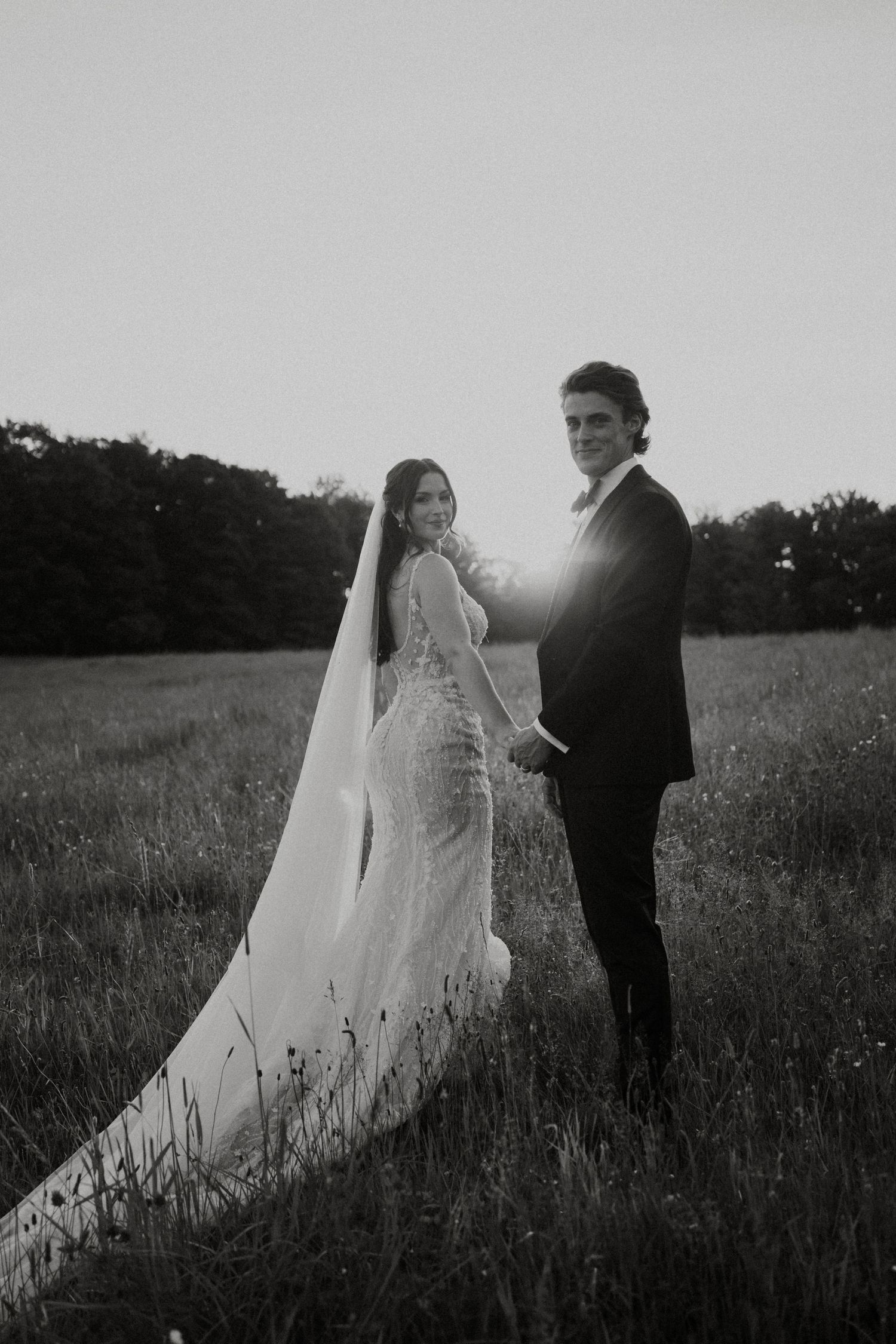 Black and white photo of bride and groom holding hands in sunlit meadow.