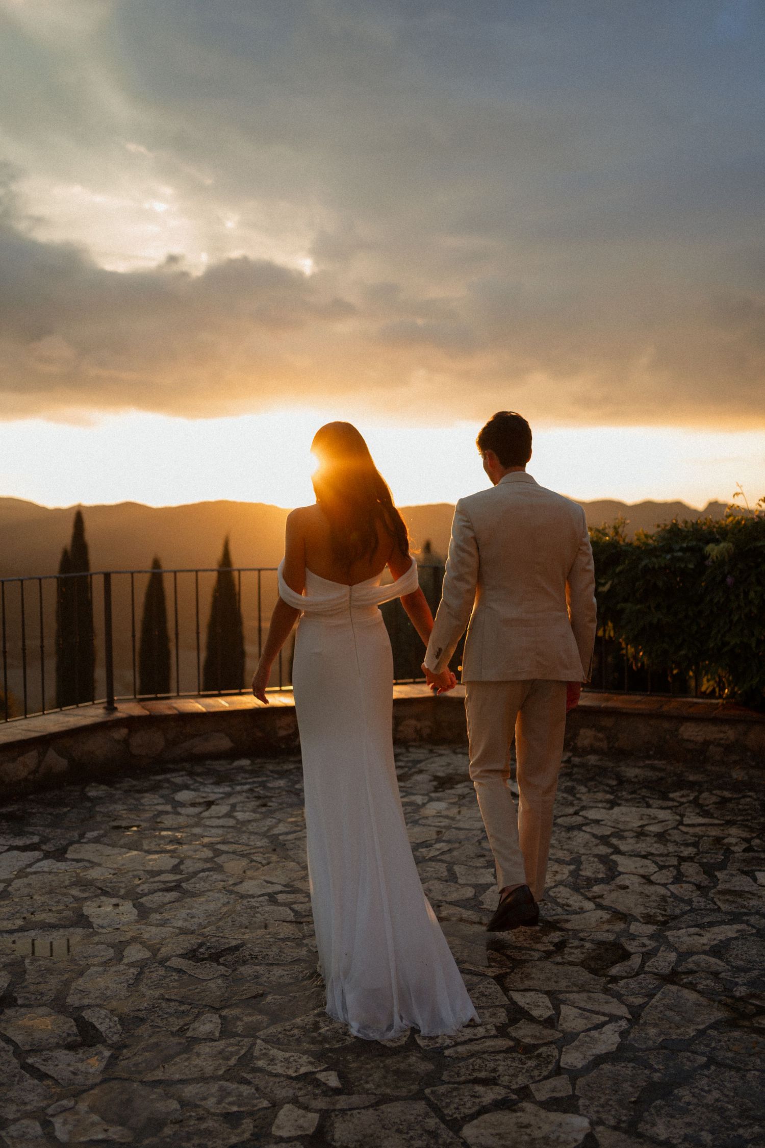 Bride and groom holding hands facing a dramatic golden sunset over Tuscan hills and cypress trees.