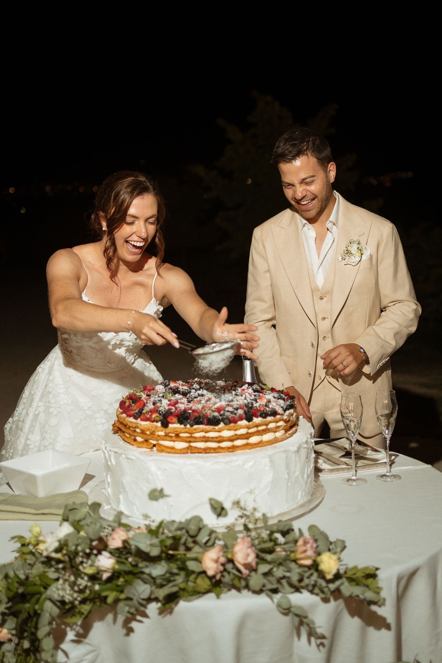 Bride and groom laughing while cutting a berry-topped wedding cake at an outdoor evening reception.
