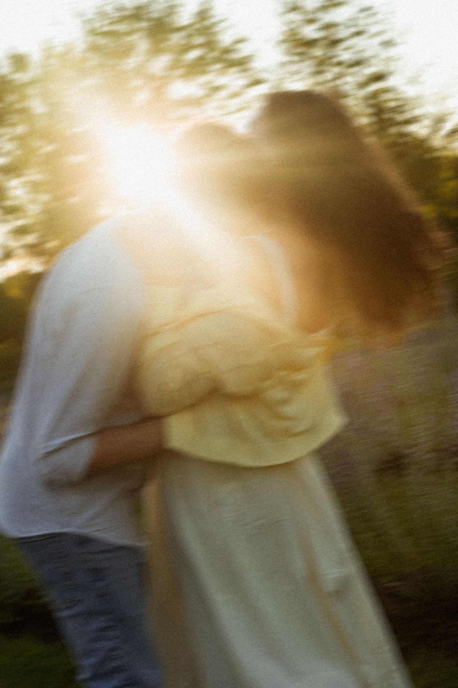 A couple embraces and kisses outdoors at golden hour, bathed in warm sunlight with a dreamy motion blur effect.