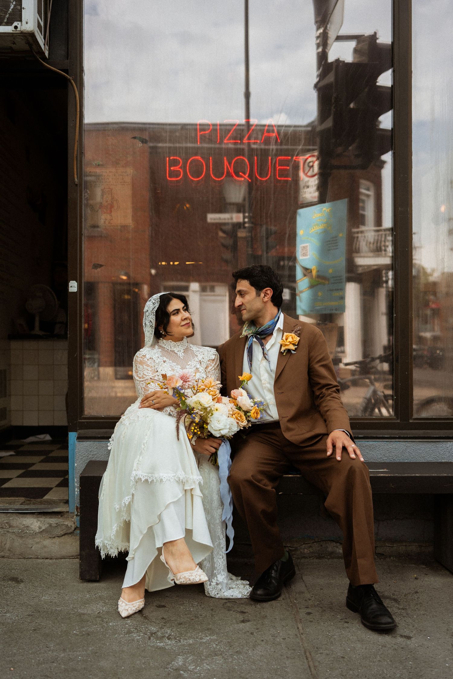 Bride and groom sit together outside a pizza shop, she in lace dress holding bouquet, he in brown suit.