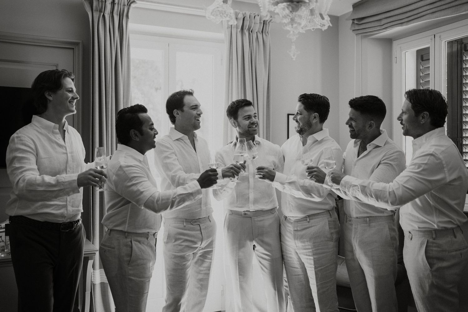 Groom and groomsmen in white shirts laughing and toasting with champagne in a bright room before wedding ceremony.