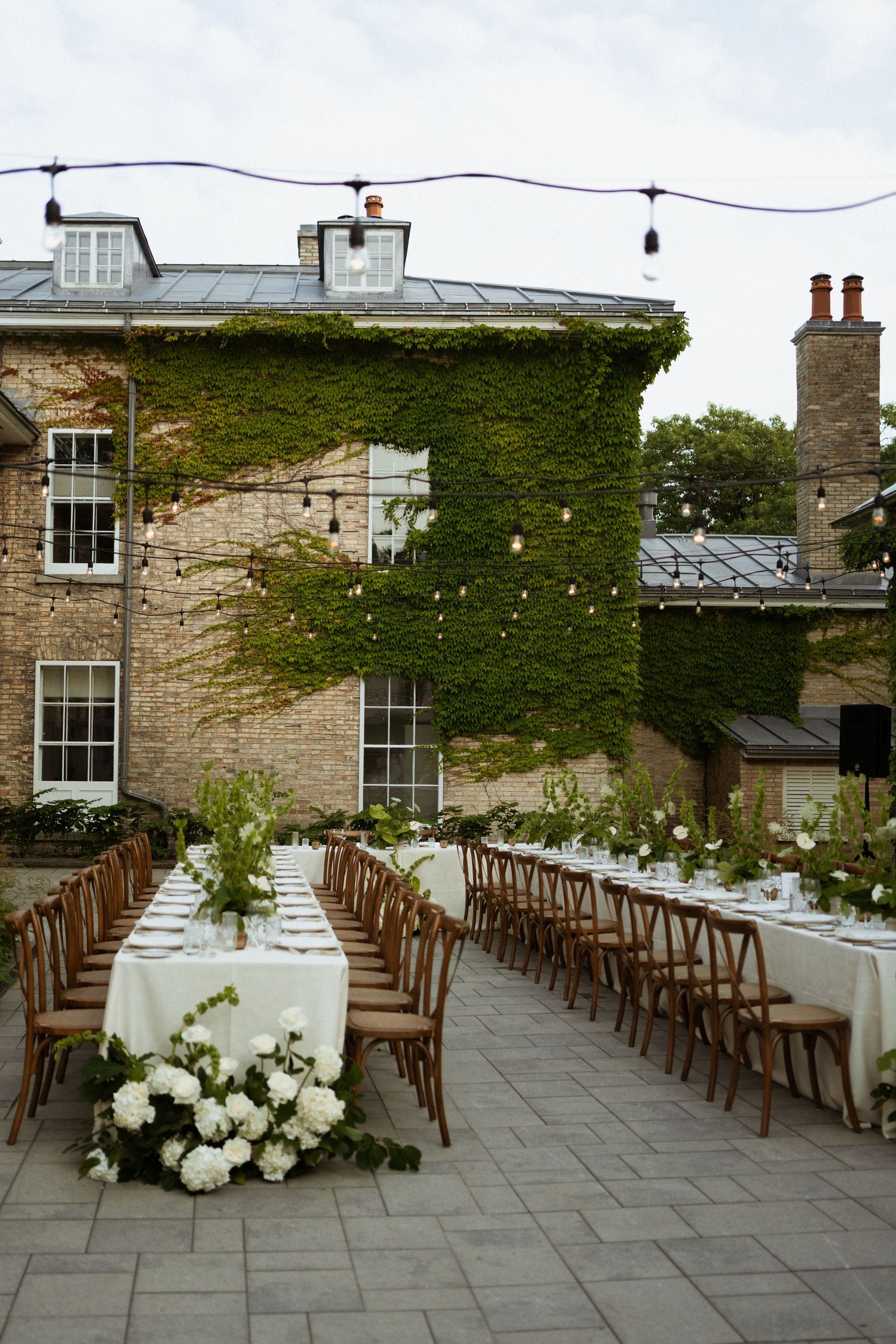 Elegant outdoor wedding reception with long tables, white florals, string lights, and ivy-covered brick building backdrop.