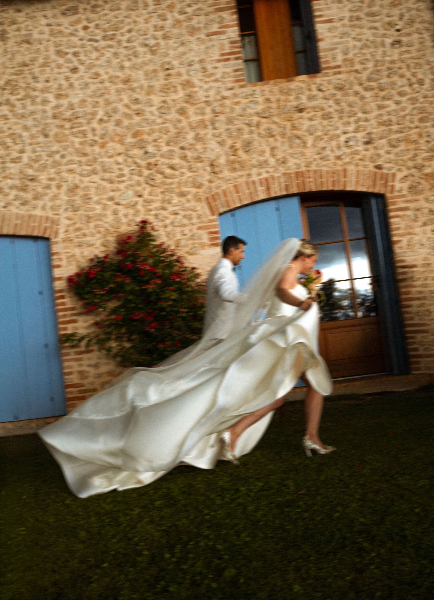 Bride in flowing white gown walks with groom along stone building with blue shutters and red flowers.