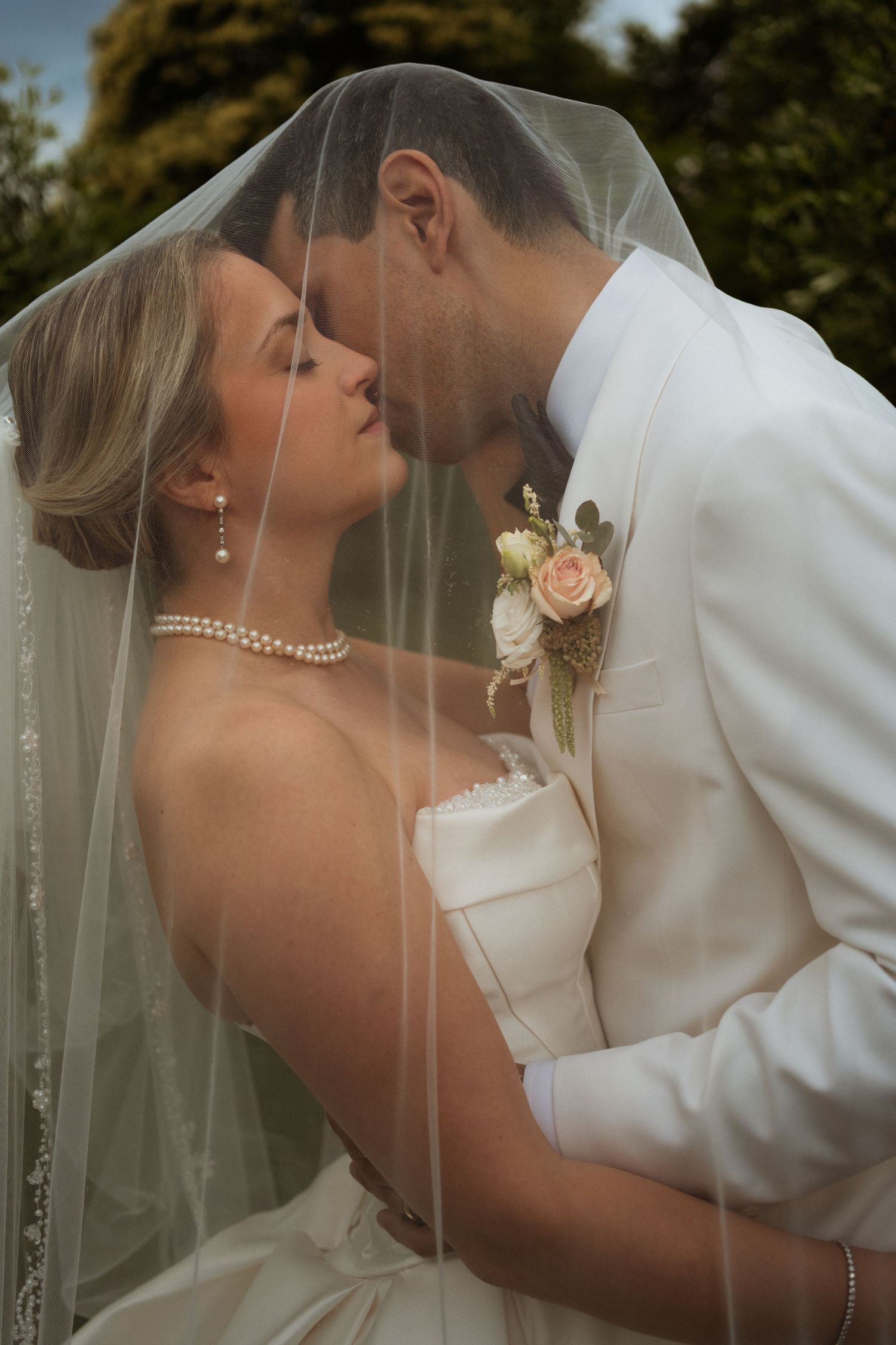 Bride and groom sharing a romantic kiss beneath a sheer wedding veil outdoors.