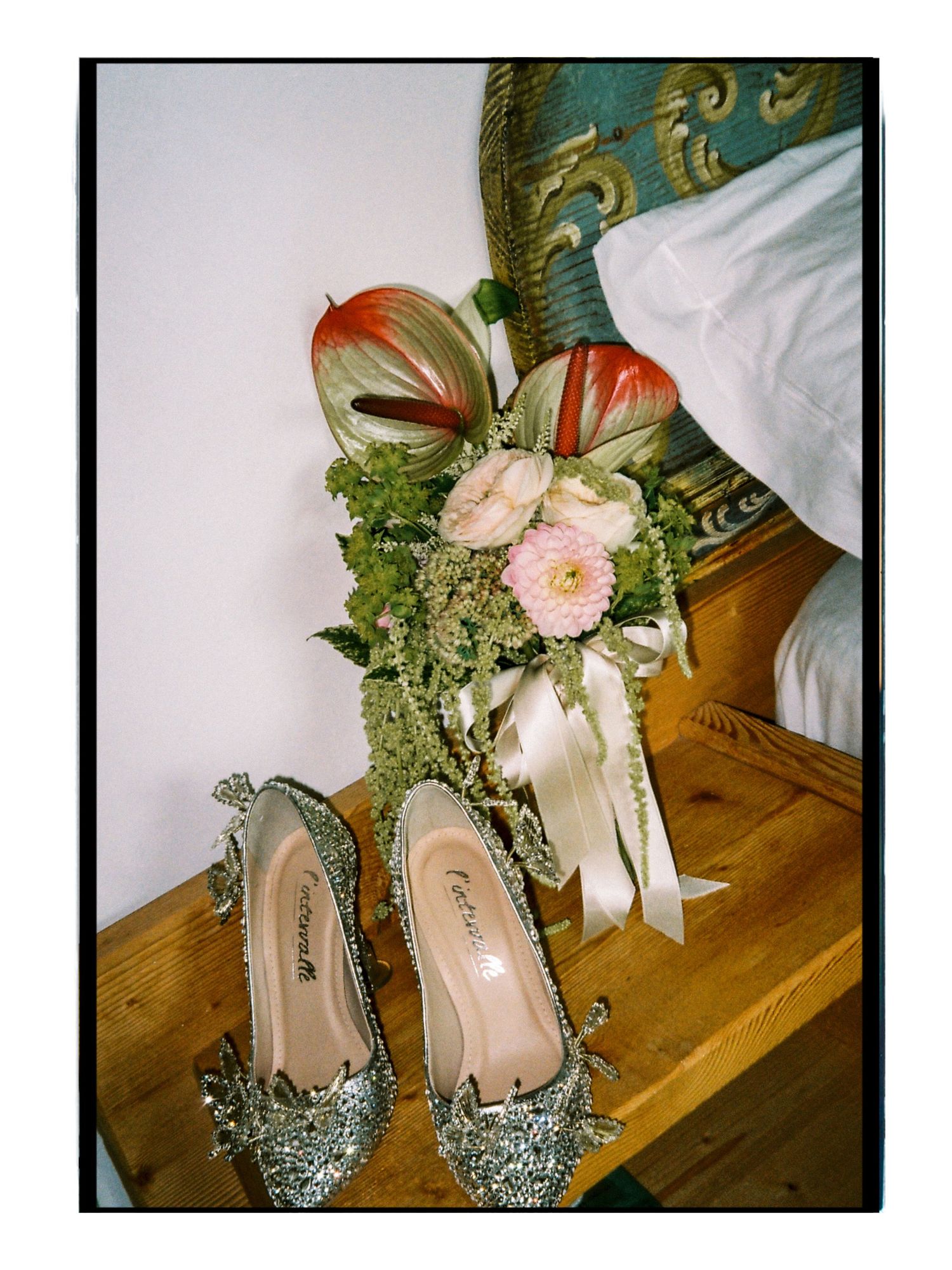 Glittery silver bridal heels beside a pink floral bouquet with ribbons on a wooden table.