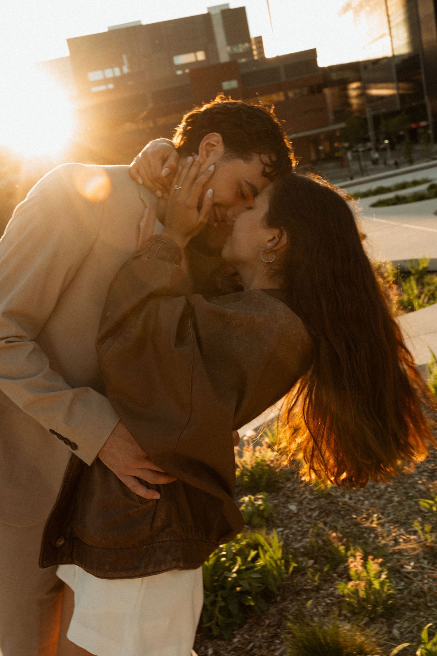 Couple sharing a romantic kiss at golden hour sunset with warm sunlight glowing behind them in an urban outdoor setting.