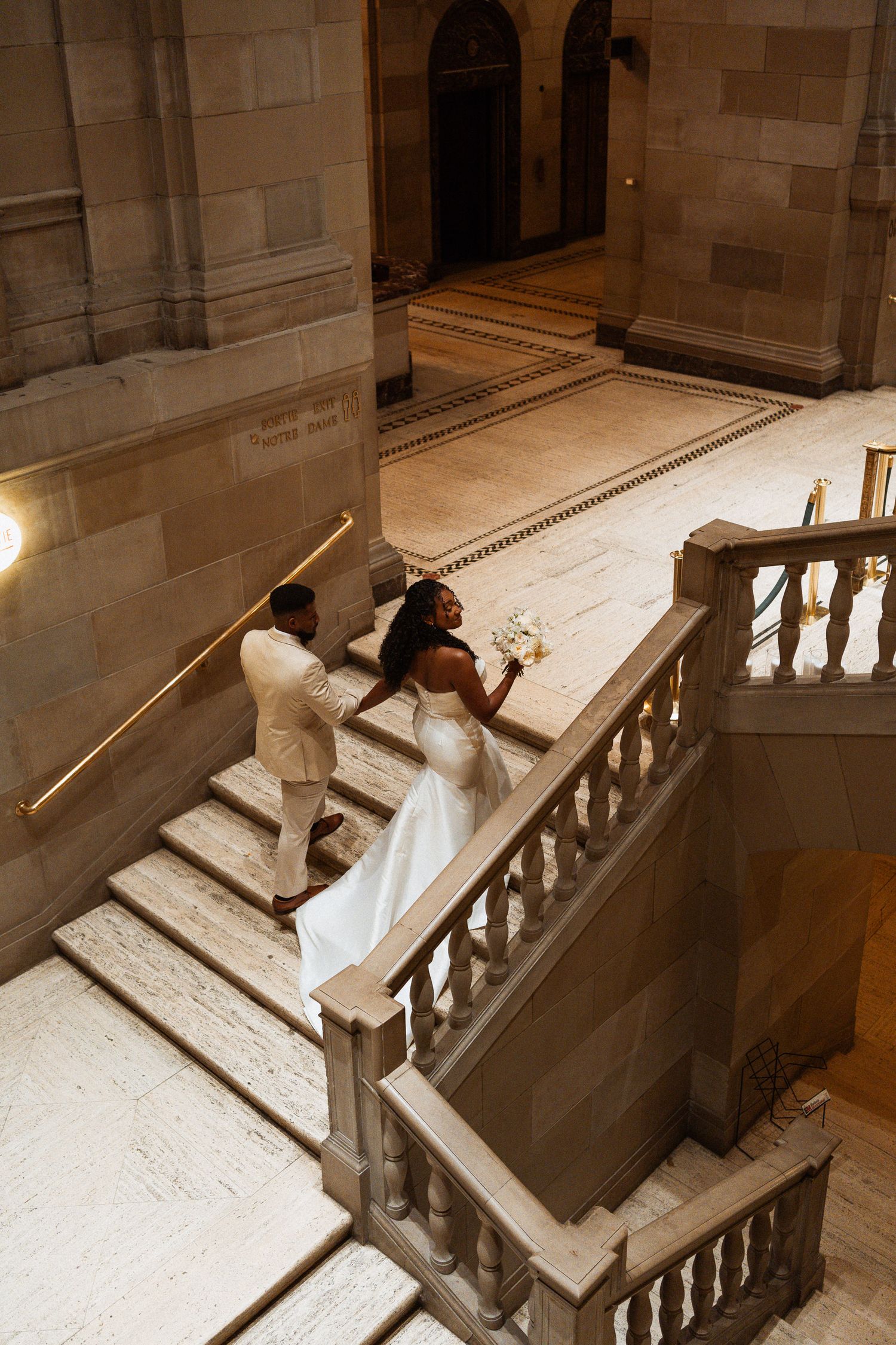 Bride and groom walking up marble staircase in a grand historic building, bride holding white bouquet.