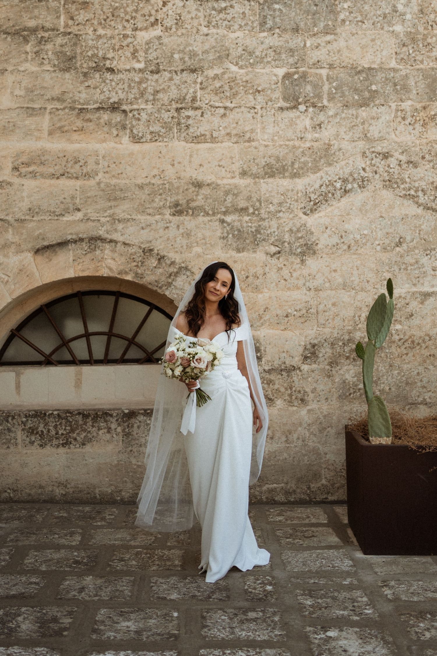 Stunning bride in white wedding dress with veil holding bouquet against rustic stone wall.