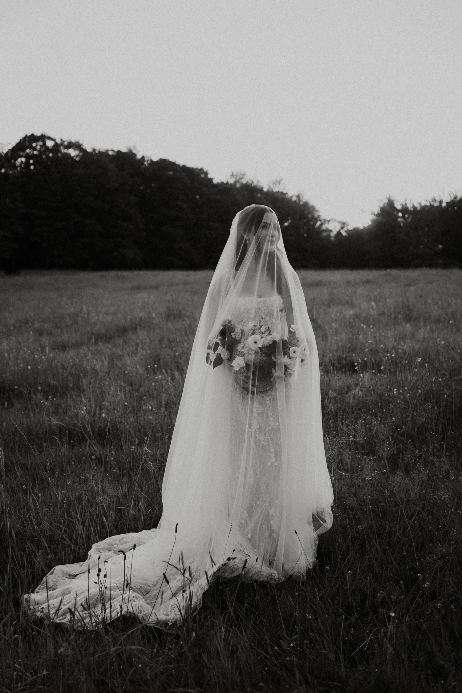 A bride in a lace gown and long veil stands alone in a misty field holding a bouquet in black and white.