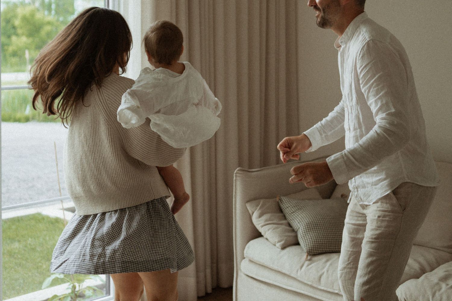 A young family with a mother holding a baby and a father dancing together in a bright, modern living room.