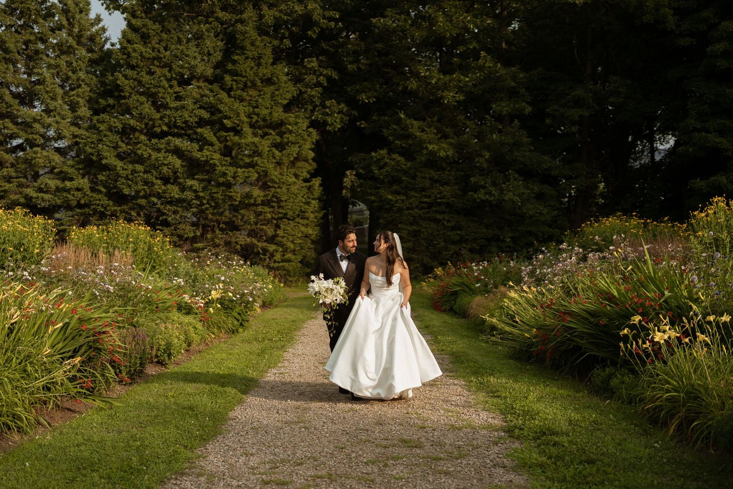 Bride and groom walking hand in hand down a lush garden path lined with colorful flowers and tall trees.