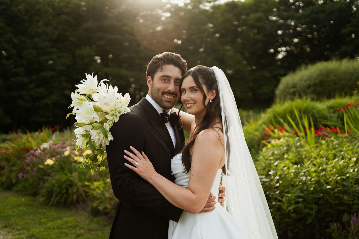 Bride and groom embrace outdoors at golden hour, bride holding white lily bouquet and wearing veil, groom in black tuxedo.