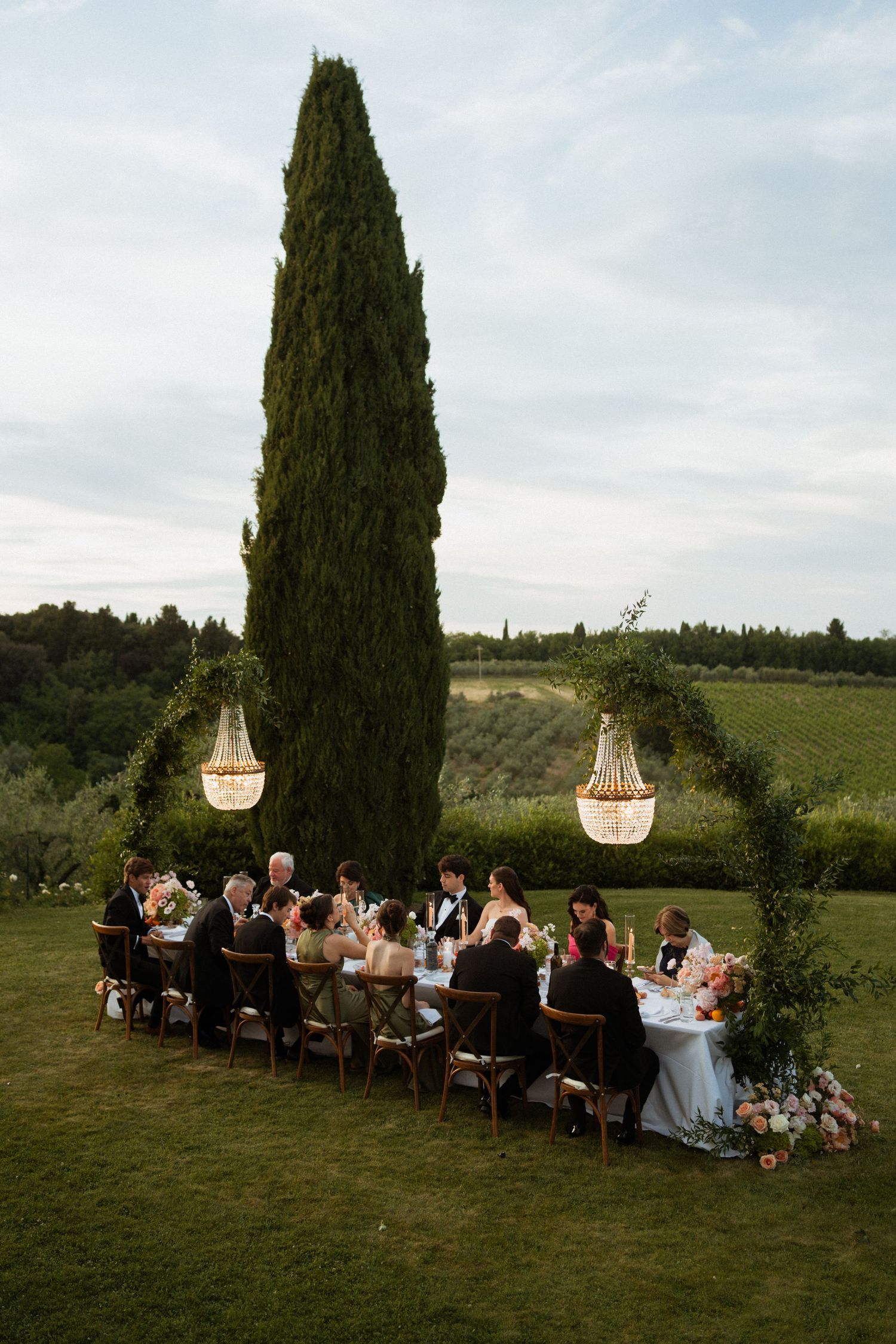 Intimate outdoor wedding reception dinner under crystal chandeliers beside tall cypress tree in Tuscany.