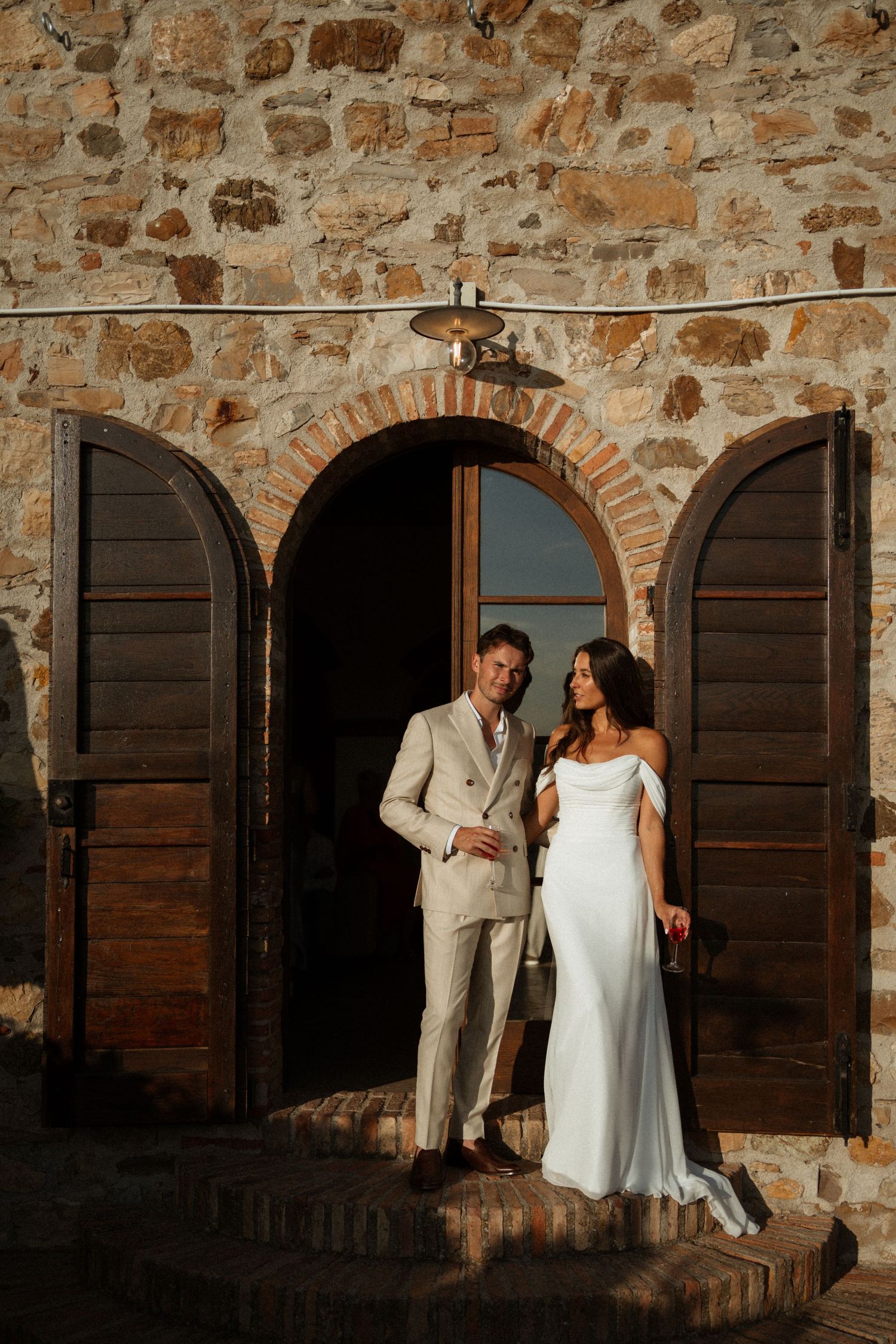 Bride and groom posing in front of rustic stone building with arched wooden doors at golden hour.