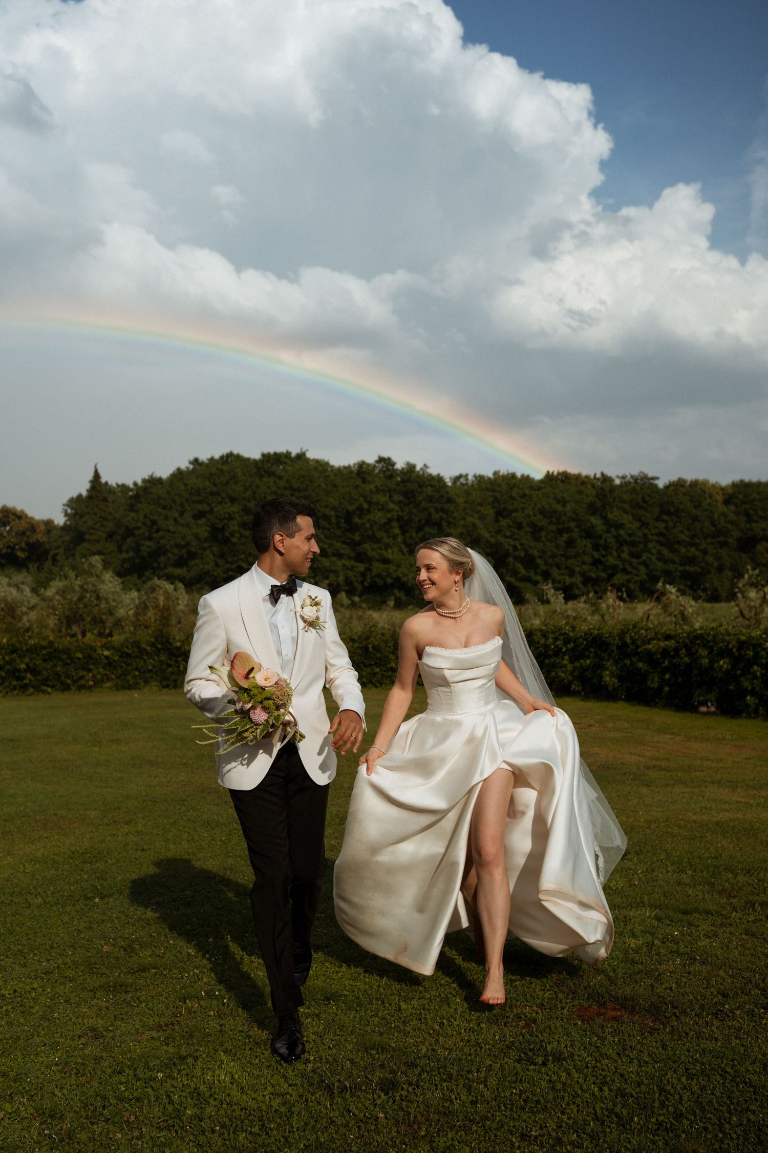 Bride and groom walking hand in hand on green lawn with a rainbow arching behind them.