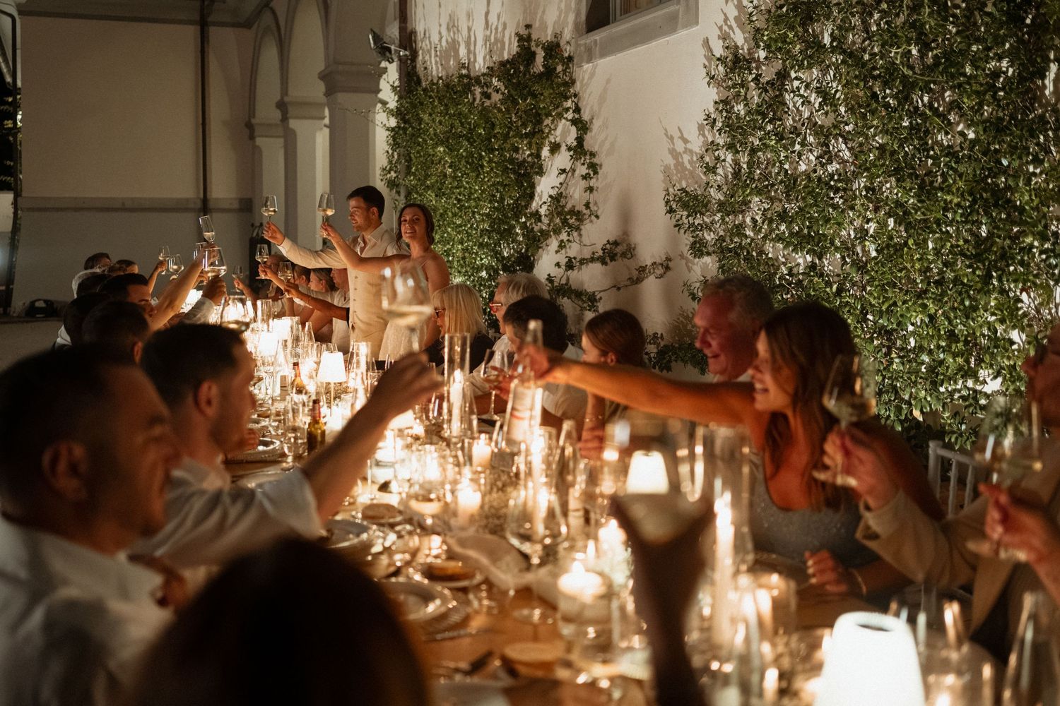 Guests raising champagne glasses in a candlelit outdoor dinner party at night with lush greenery backdrop.