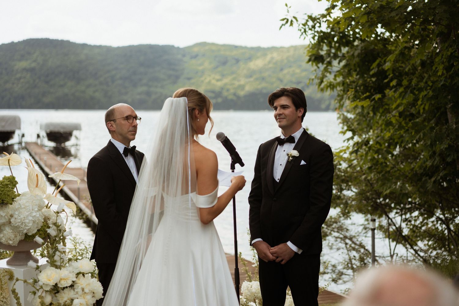 Bride and groom exchange vows outdoors by a lake with lush green hills, officiant beside them, white floral arrangements nearby.