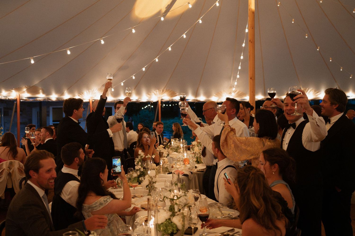 Guests raise glasses in a toast under a elegant tent at a candlelit wedding reception dinner party.