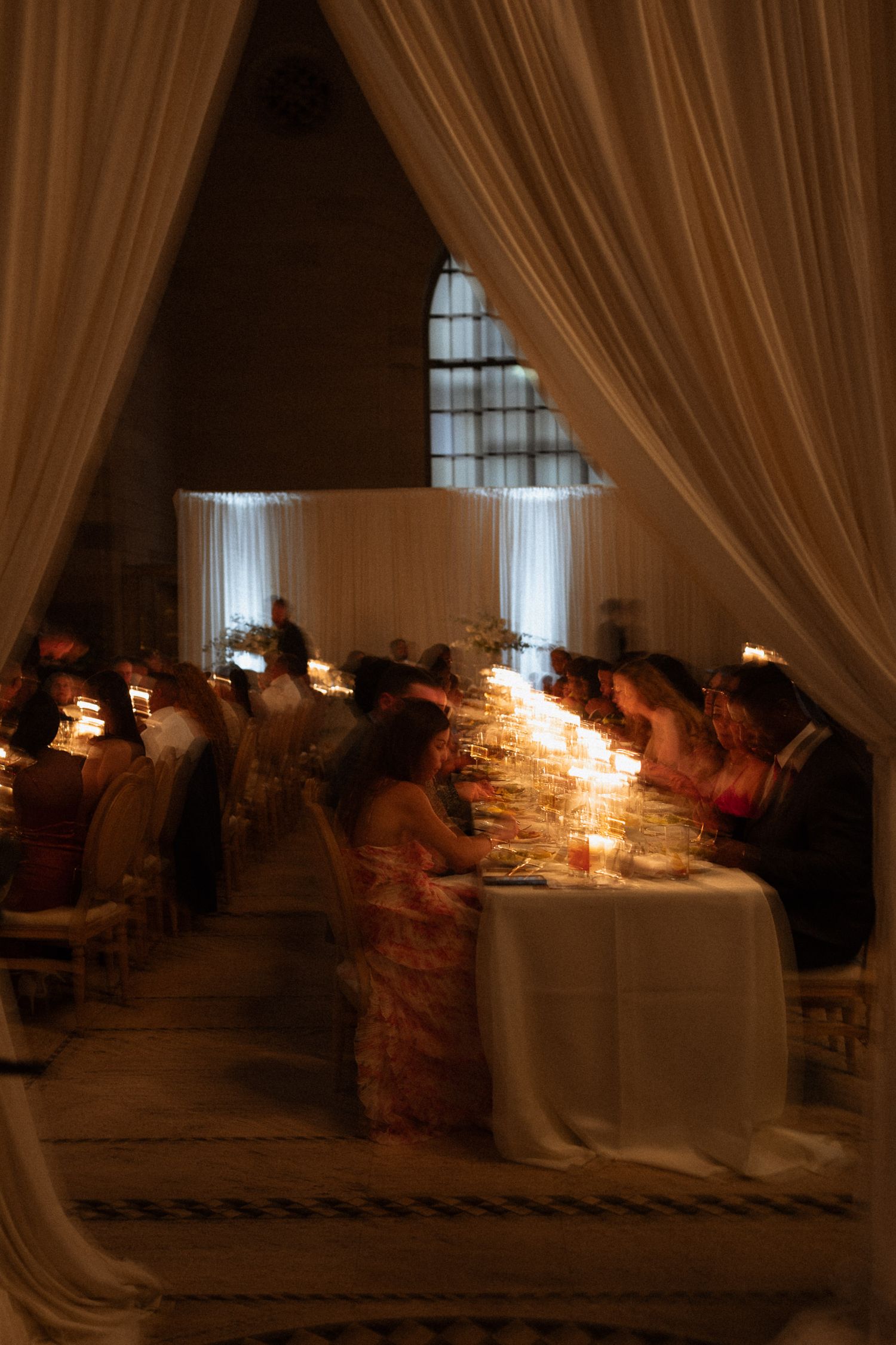 Elegant candlelit wedding reception with guests seated at long tables, draped white curtains framing the romantic dimly lit venue.