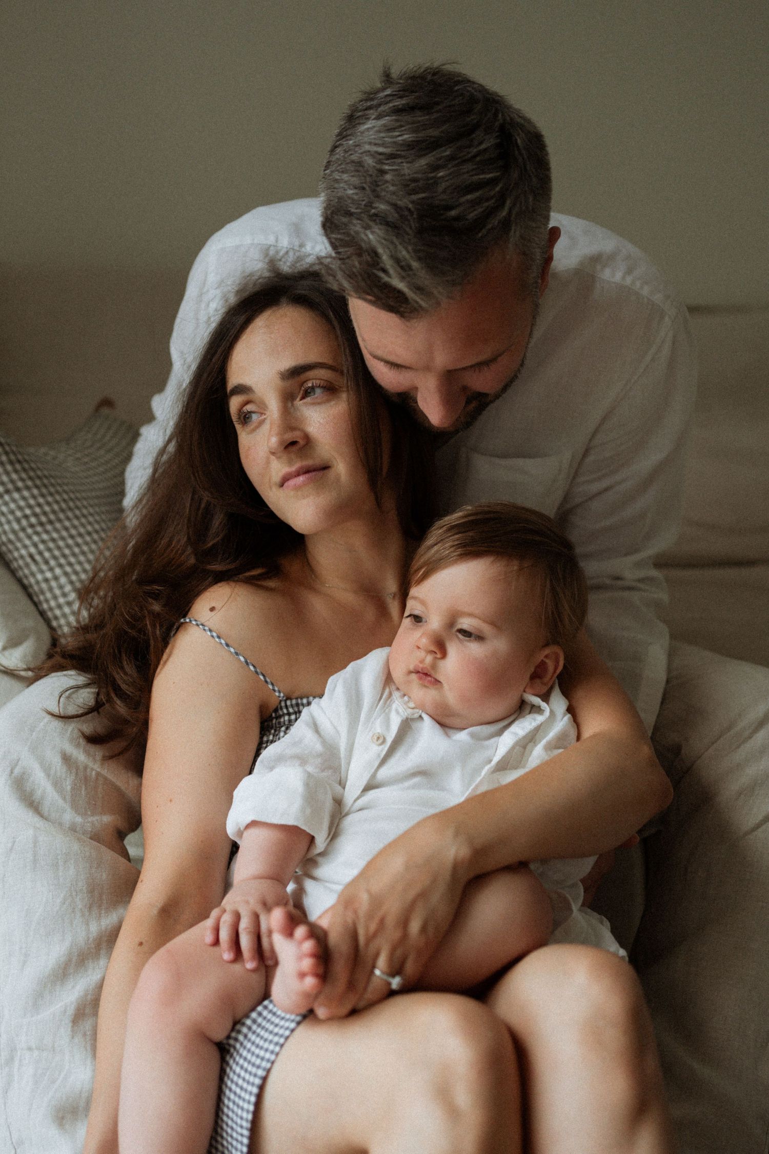 A family portrait of a mother, father, and baby sitting together on a bed, with the father lovingly leaning over them.