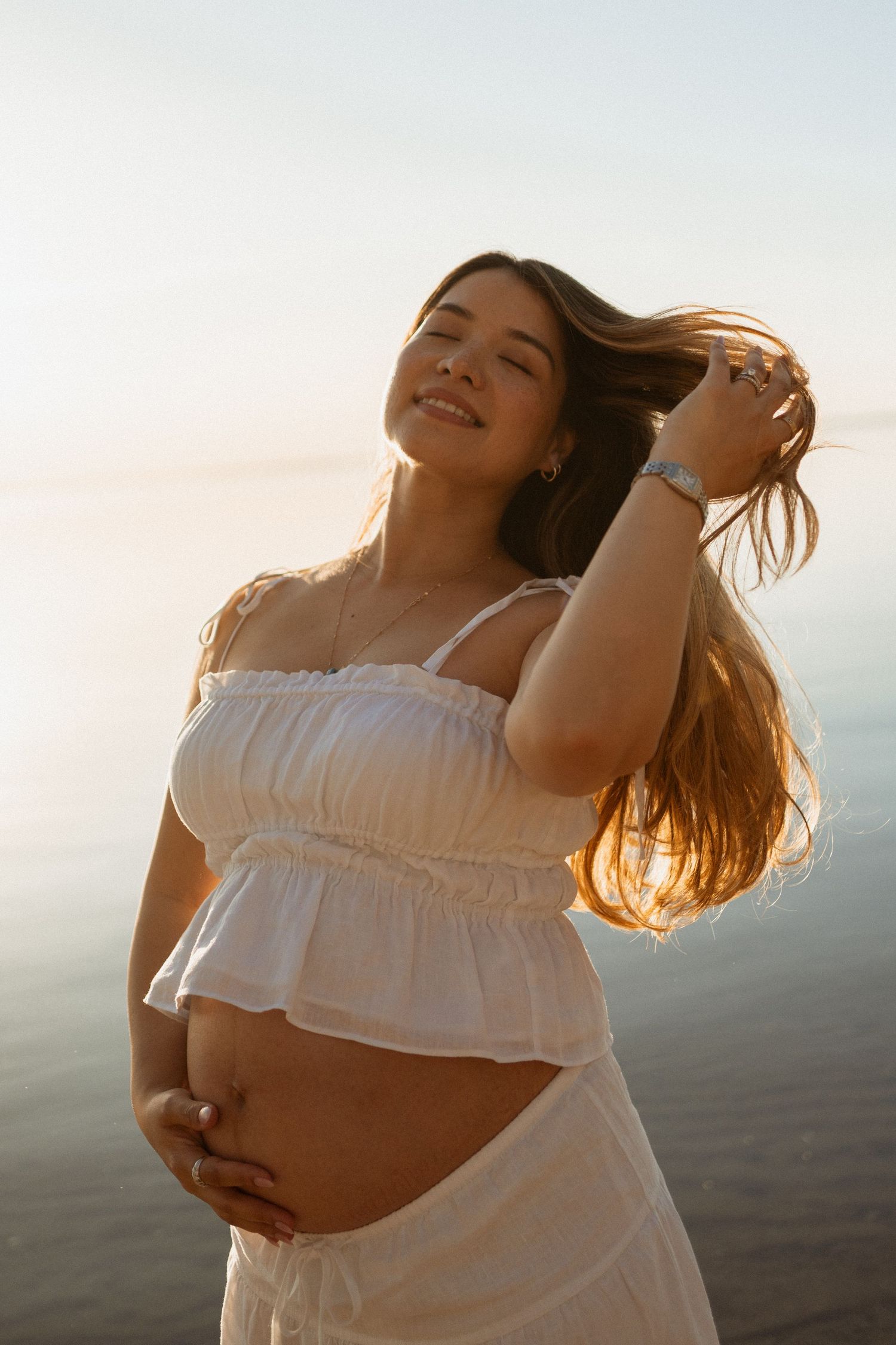 Smiling pregnant woman in white crop top holding her baby bump at sunset by the water.