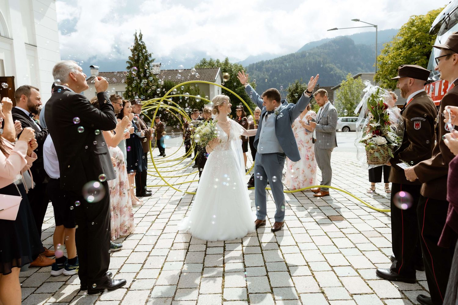 Newlyweds walk through bubble send-off with cheering guests and mountain backdrop after wedding.