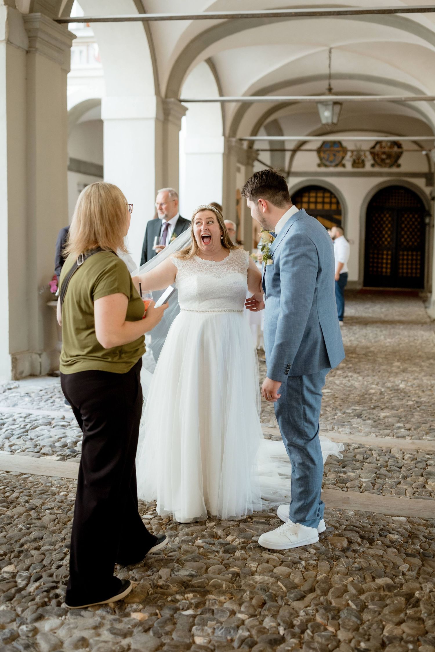 Joyful bride in white gown laughing with guests under arched columns at a wedding venue.