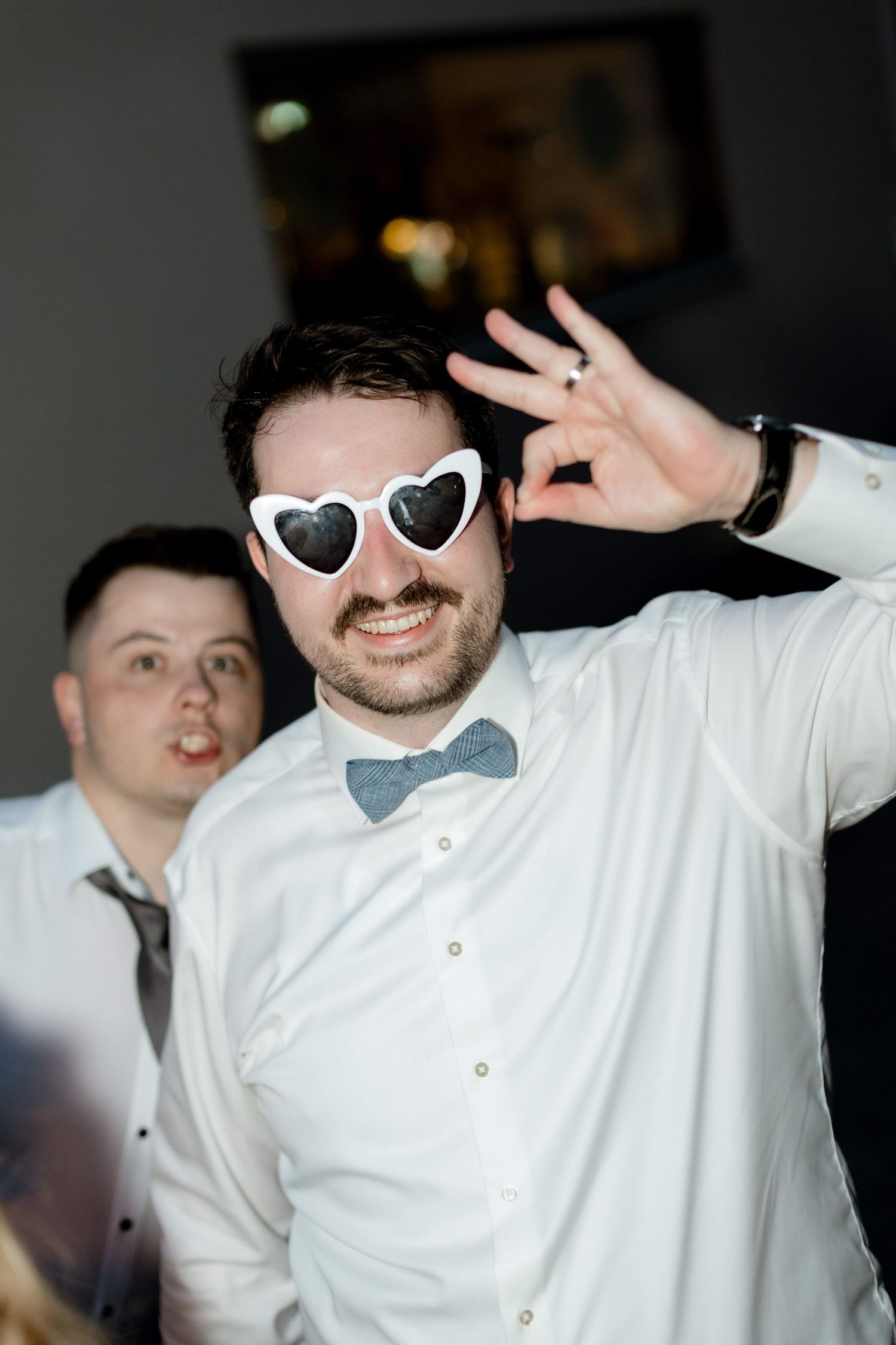 Grinning man in white shirt and bow tie wearing heart-shaped sunglasses at a wedding party.