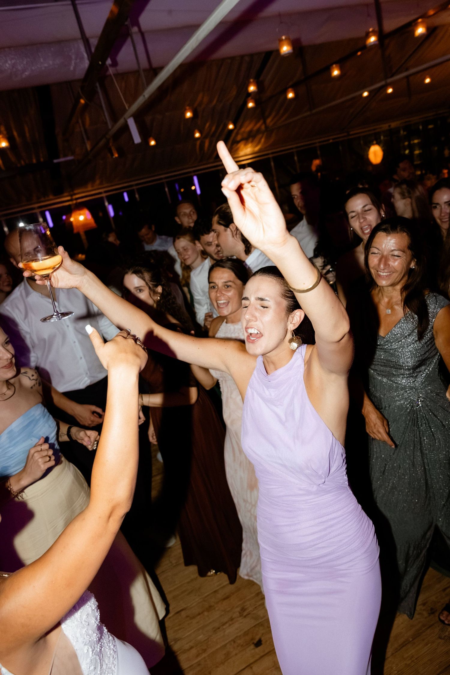 Woman in lavender dress dancing with raised arms among wedding guests on dance floor.