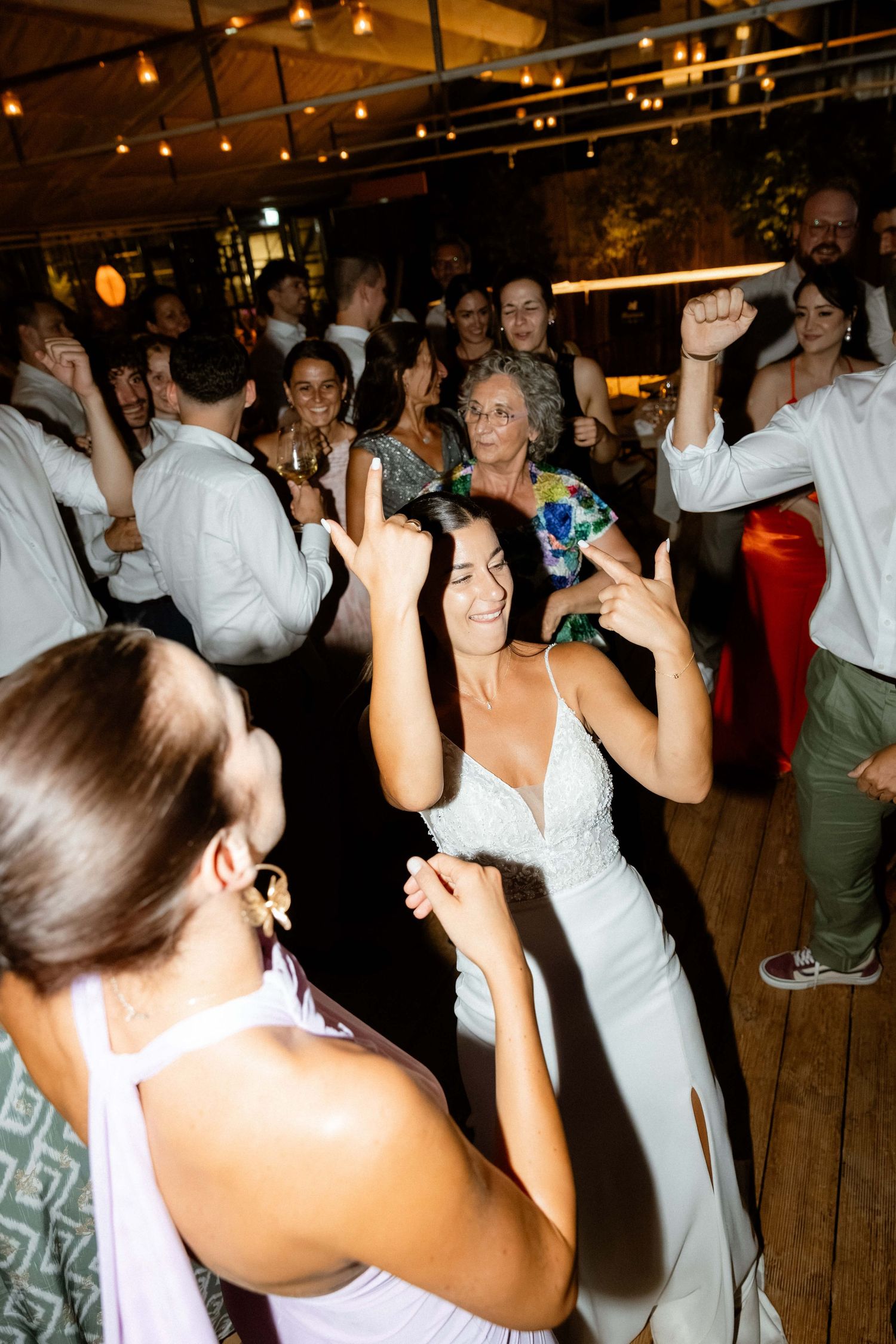 Bride in white gown dancing joyfully with guests at wedding reception under warm string lights.