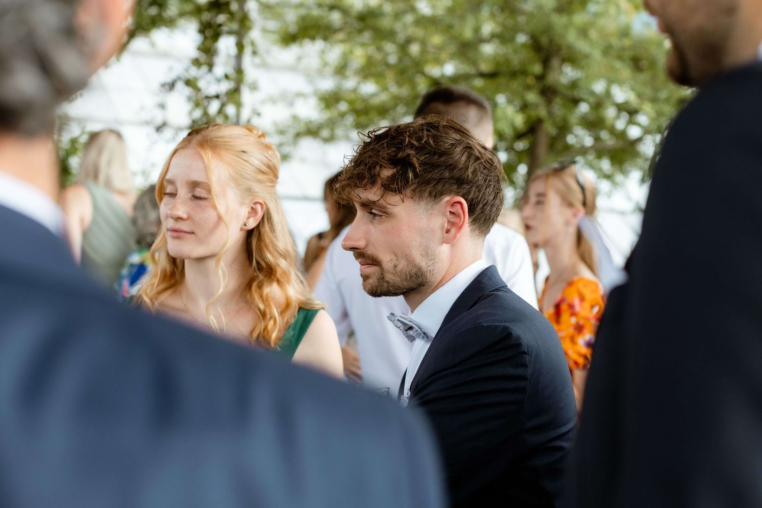 Young wedding guests in formal attire socializing outdoors under green trees at a summer celebration.