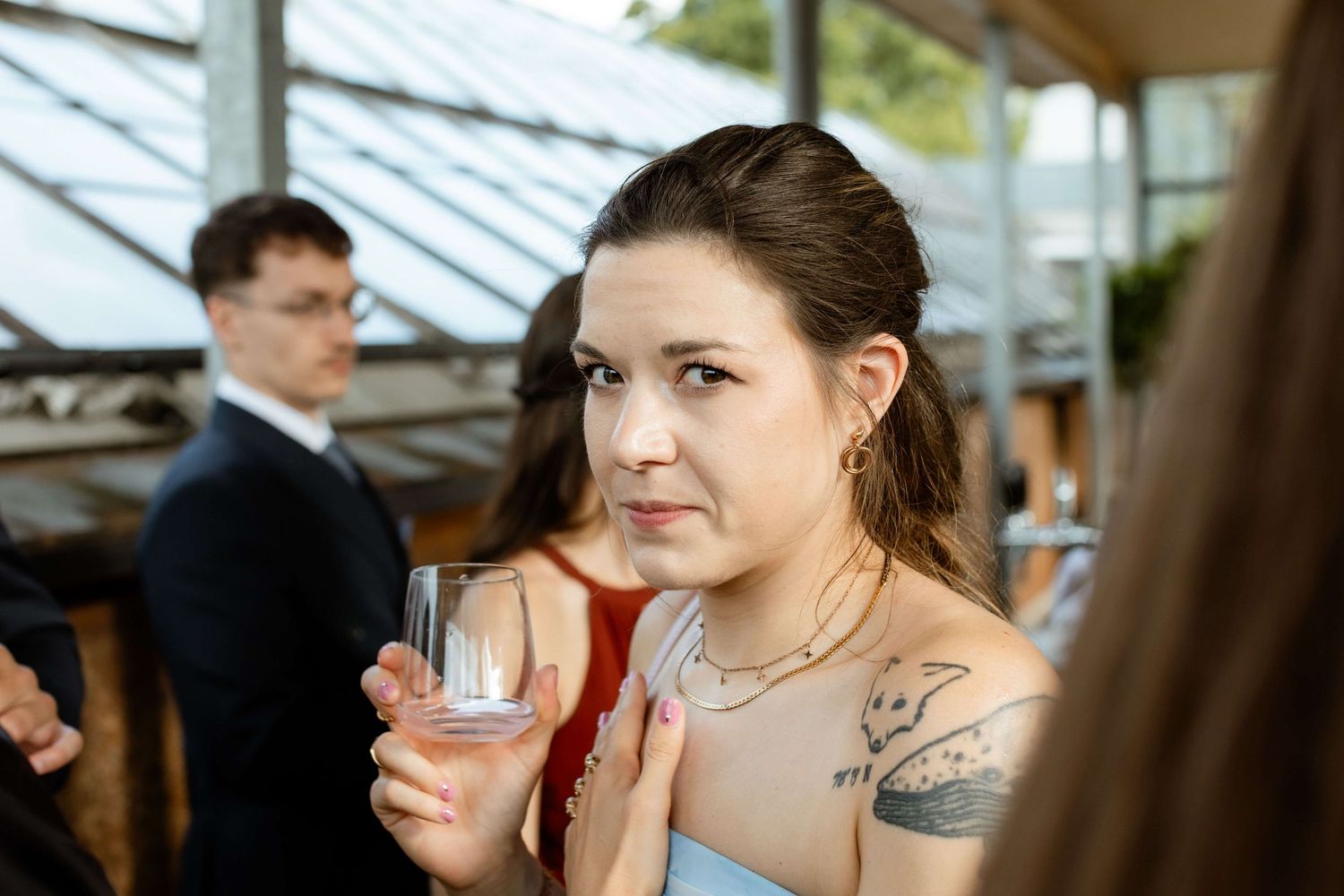 Young tattooed woman holding a glass at a wedding reception wearing a strapless dress and layered necklaces.