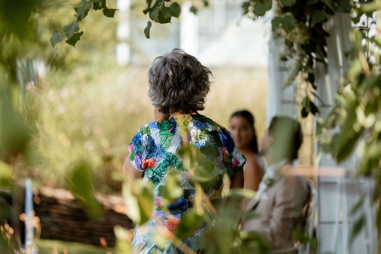 Guest in colorful floral dress viewed from behind at a lush green outdoor wedding ceremony.