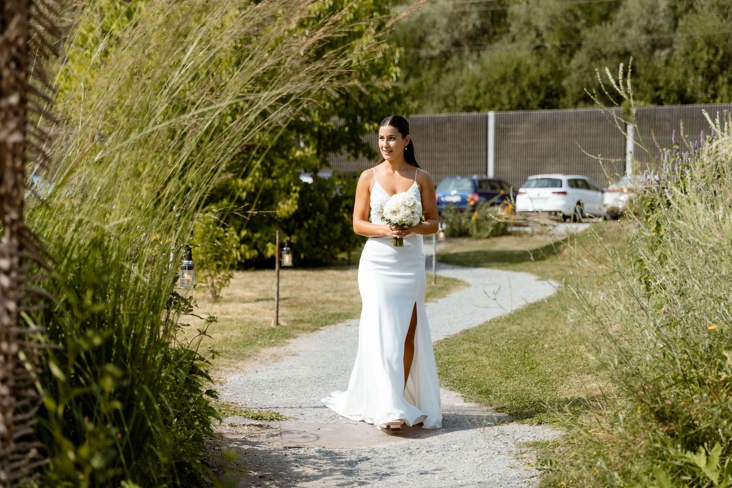 Bride in elegant white gown holding bouquet walking along a garden path on wedding day.