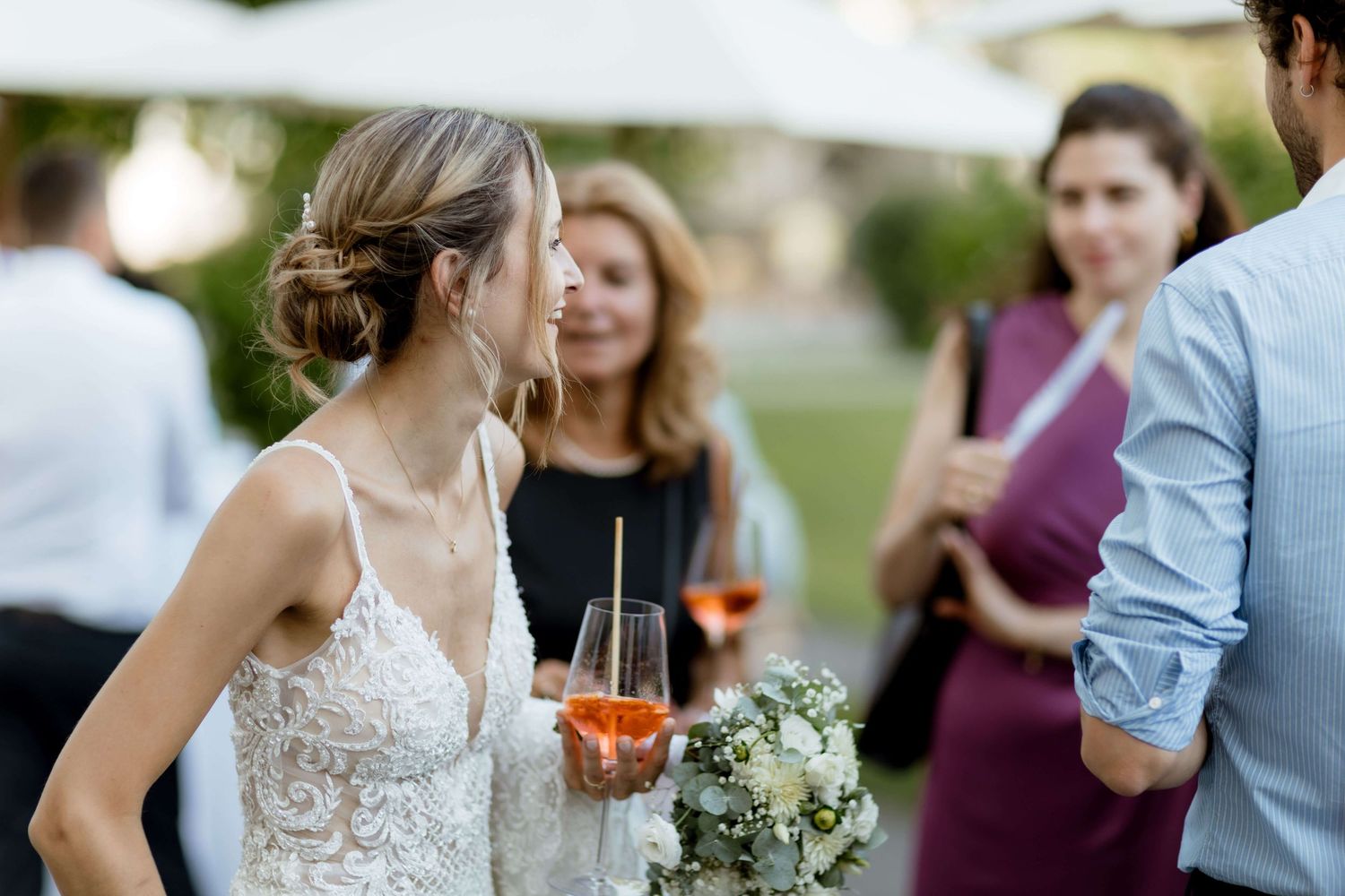 Bride in lace gown holds bouquet and rosé wine while chatting with guests at reception.