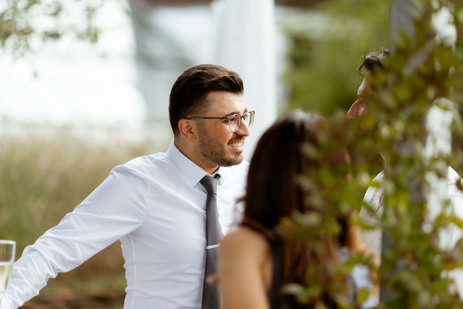 A well-dressed man in glasses and a dark tie sits relaxed at an outdoor wedding reception surrounded by greenery.