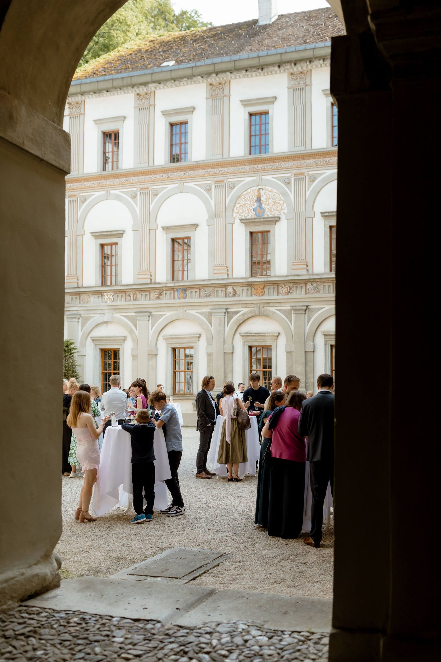 Wedding guests mingling in a sunlit courtyard of a grand Renaissance-style palace during a reception.