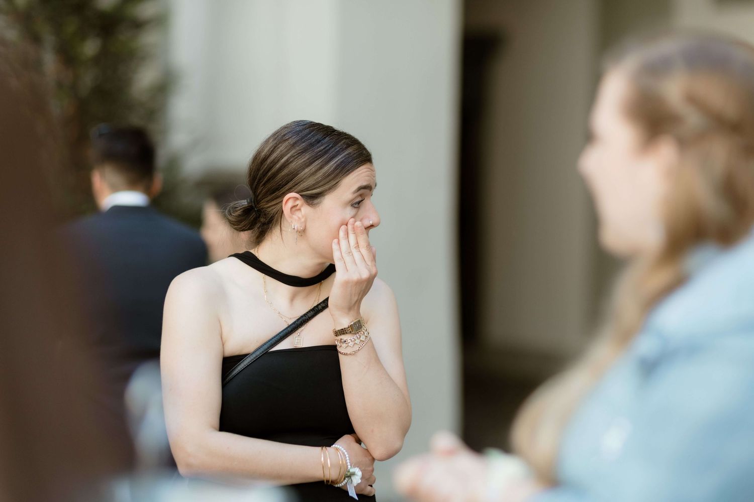 Young woman in black dress covering mouth while chatting with friend at outdoor wedding event.