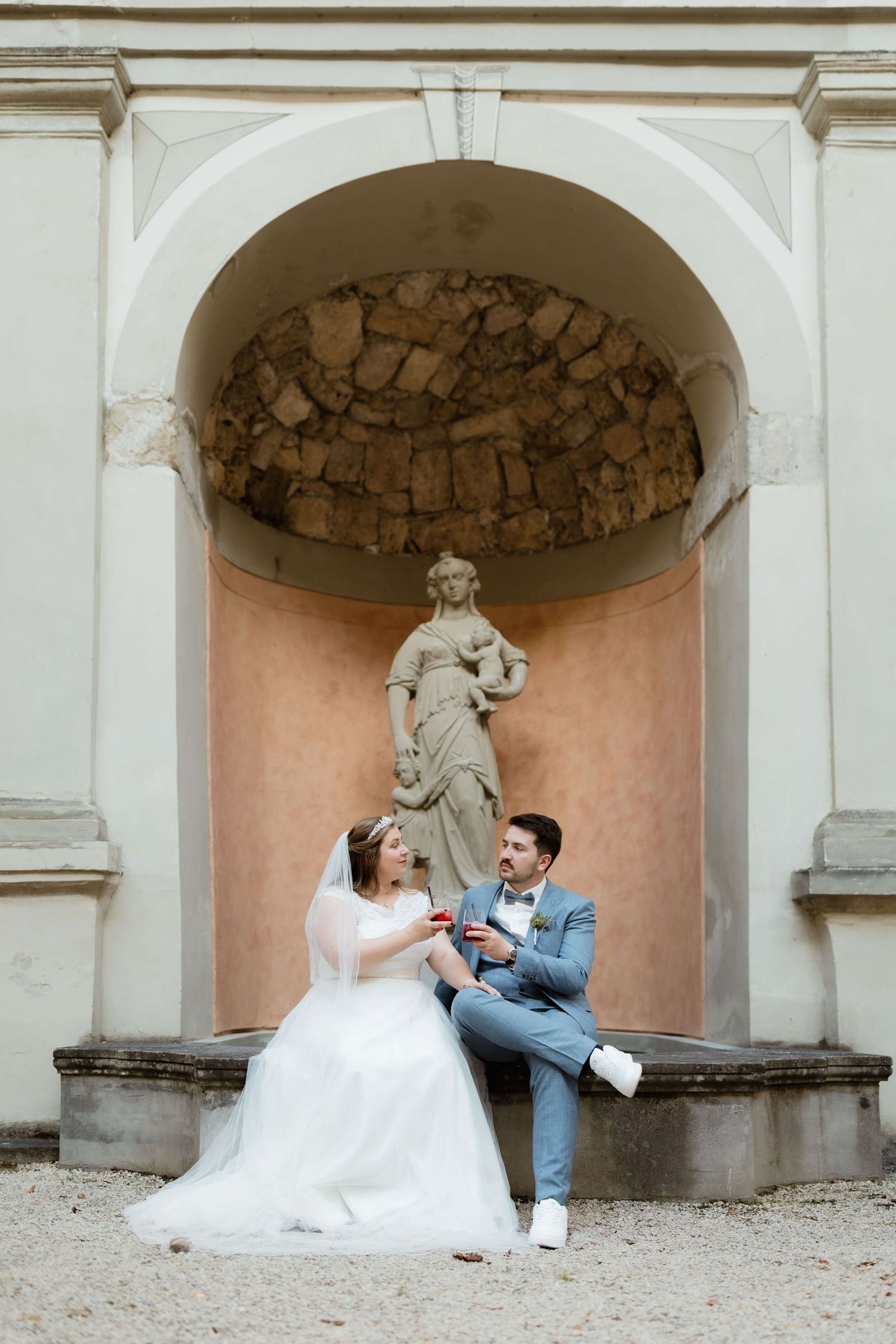 Bride and groom sit together in front of a classical stone arch with a female statue, exchanging rings outdoors.