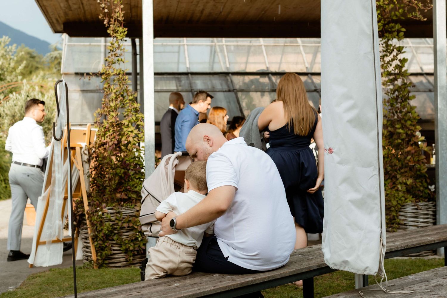 Wedding guests relax on outdoor benches near a modern greenhouse venue surrounded by lush greenery.