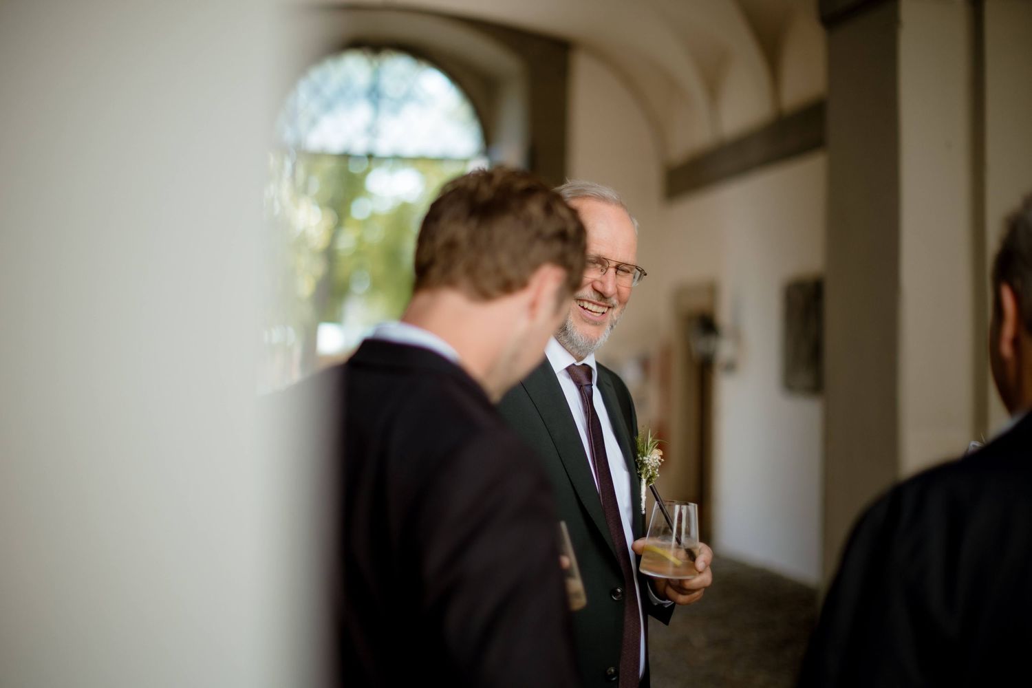Smiling older man in a dark suit holding a drink while conversing with guests at a wedding reception.