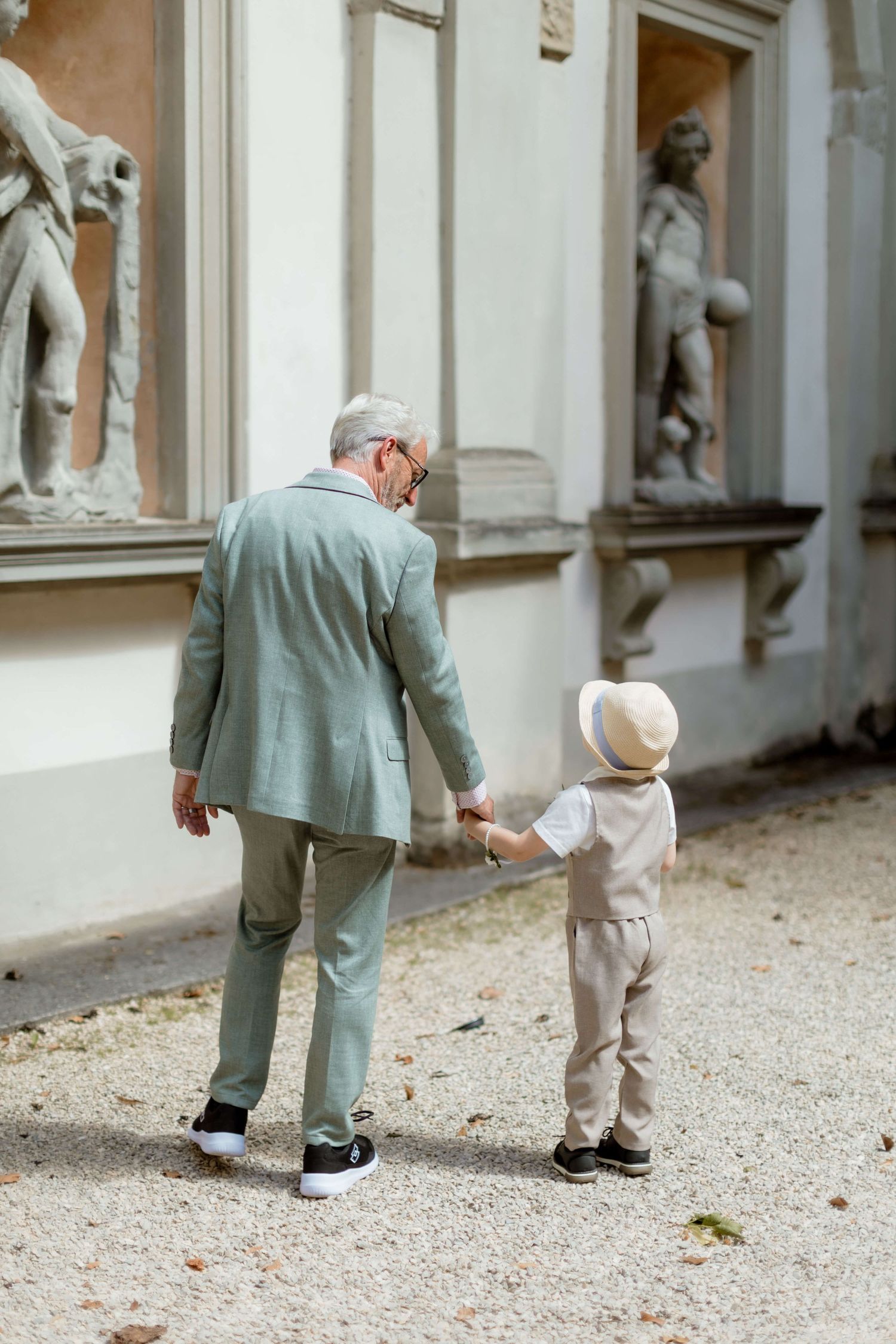 Elderly man in a sage suit holding hands with a young boy near classical sculptures at a wedding venue.