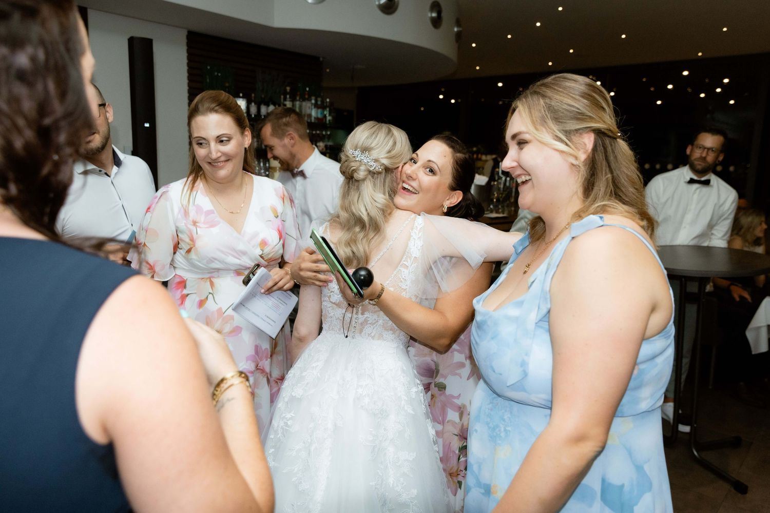 A joyful bride in a white dress celebrates with bridesmaids holding a champagne bottle at a wedding party.