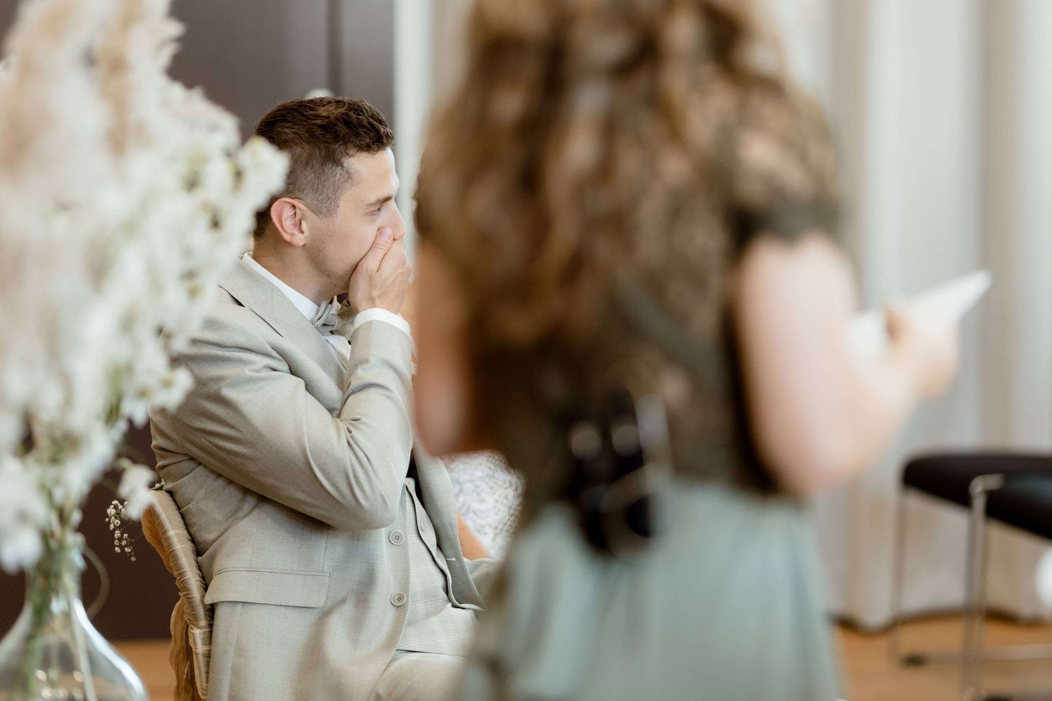 Emotional groom in tan suit covers mouth with hand during heartfelt wedding ceremony speech indoors.